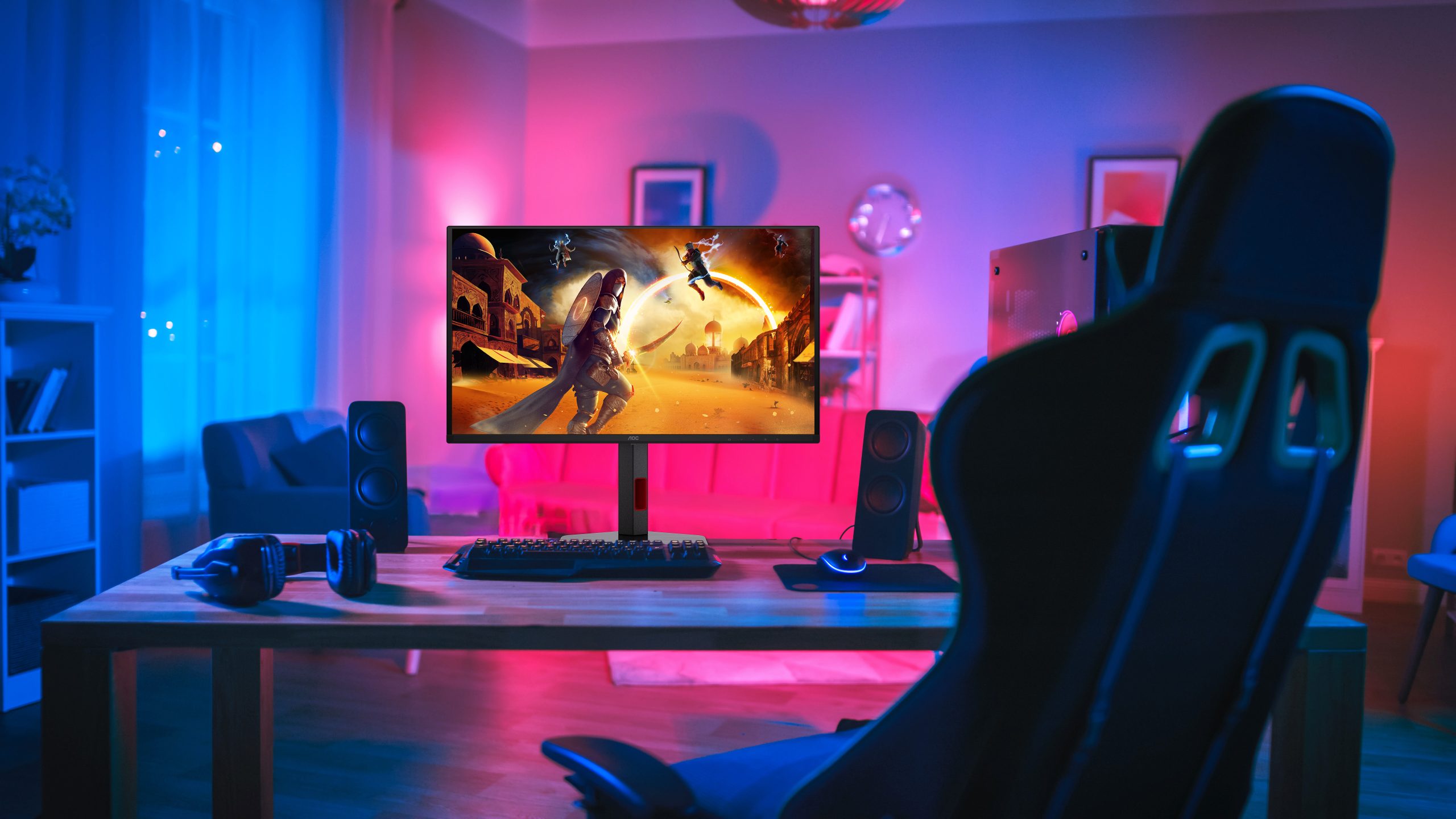 AOC gaming monitor displaying a fantasy game scene on a desk with speakers, headphones, and a gaming chair in a dimly lit room.