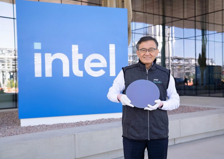 Man in Intel Foundry vest holding a silicon wafer in front of an Intel sign outdoors.