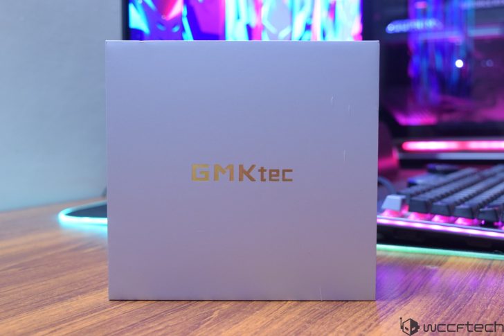 A white box with the gold logo 'GMKtec' is on a desk in front of a gaming setup with colorful lighting.