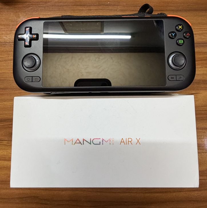 A handheld gaming console with 'MANGMI AIR X' branding on a white box, featuring two joysticks and gaming controls, is placed on a wooden surface.