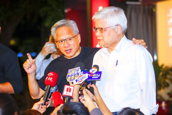 Two men at a press conference surrounded by microphones, one speaking, another listening attentively.