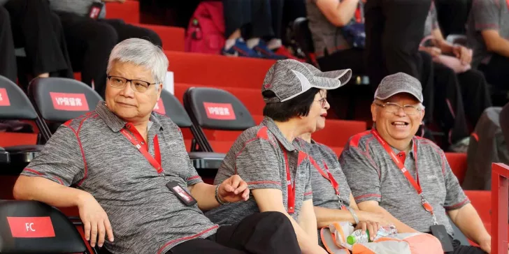 Three people in matching gray Synnex shirts and hats sit on a red bench at an event, with two visibly smiling and wearing lanyards.