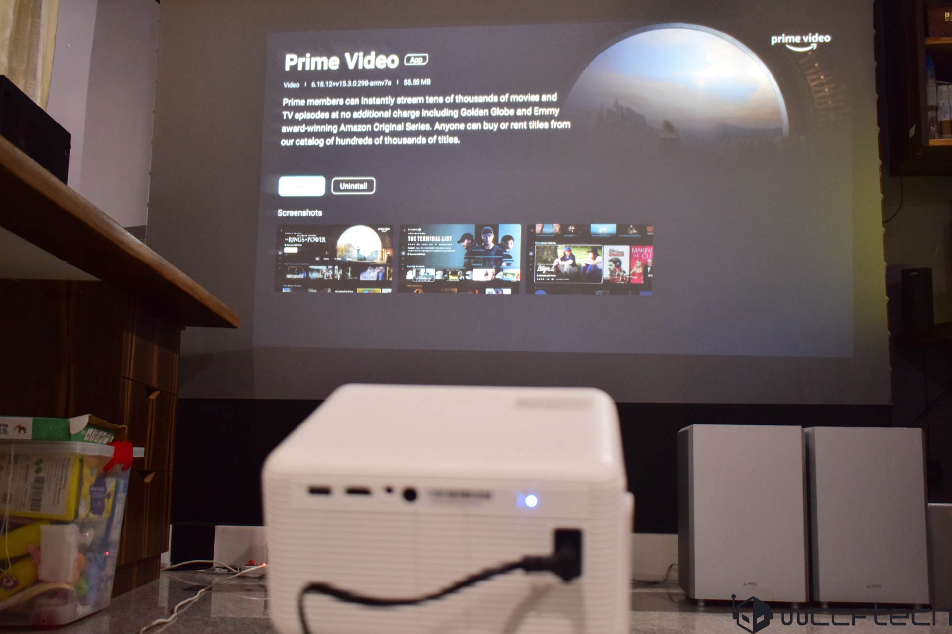 An unbranded projector displays the 'Prime Video App' page on a screen in a room, highlighting the ability to stream movies and TV episodes.