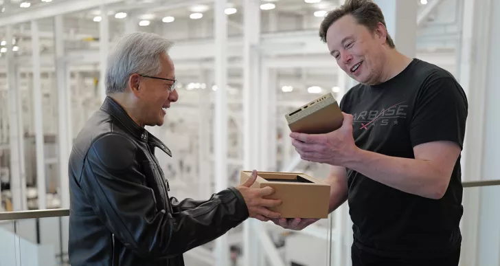 Two men smiling and interacting over a small unbranded device in a cardboard box, with one wearing a black leather jacket and the other a T-shirt with 'STARBASE' visible.