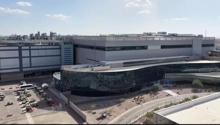 Industrial facility with a large curved building, parking lot filled with small vehicles, and visible equipment on rooftops.