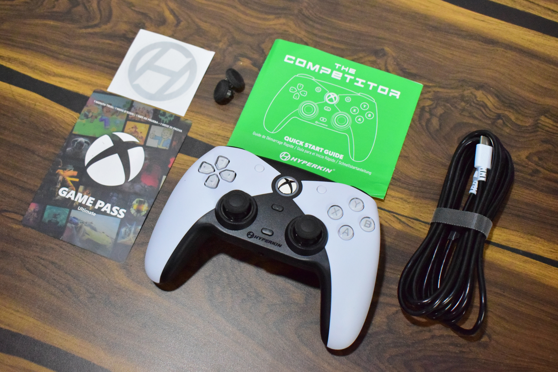 A Hyperkin The Competitor wired controller for Xbox is displayed alongside a green quick start guide, an Xbox Game Pass Ultimate card, and a connecting cable on a wooden surface.