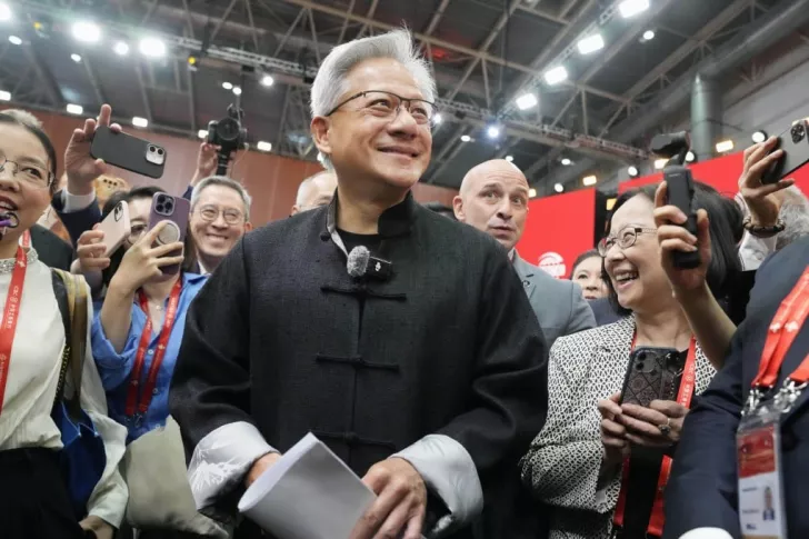 A group of people, including a smiling central figure in a black outfit, surrounded by others with smartphones at a tech event with red banners in the background.