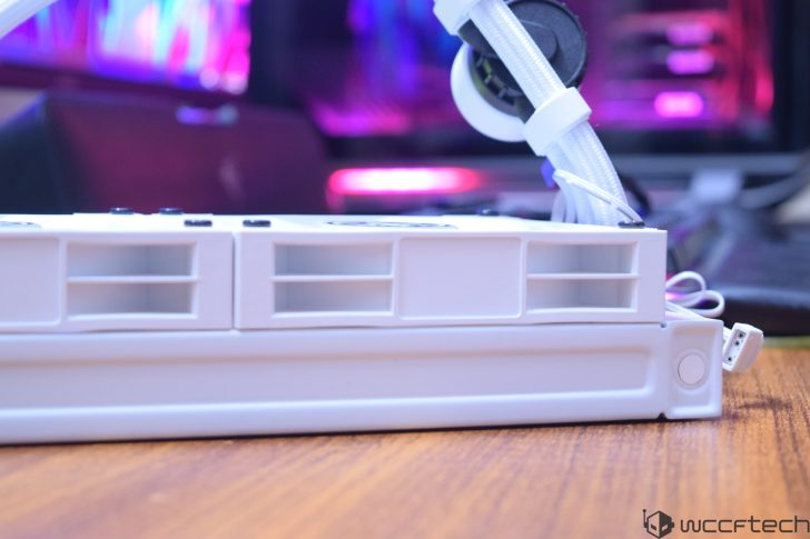 A close-up of a white PC radiator with attached tubes, placed on a wooden surface with colorful LED lights in the background.