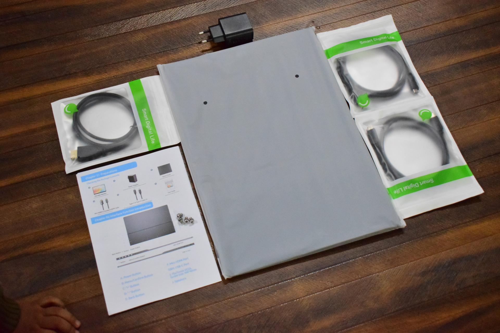 Unbranded monitor accessories and setup guide are laid out on a wooden floor, including cables in 'Smart Digital Life' packaging and a power adapter.