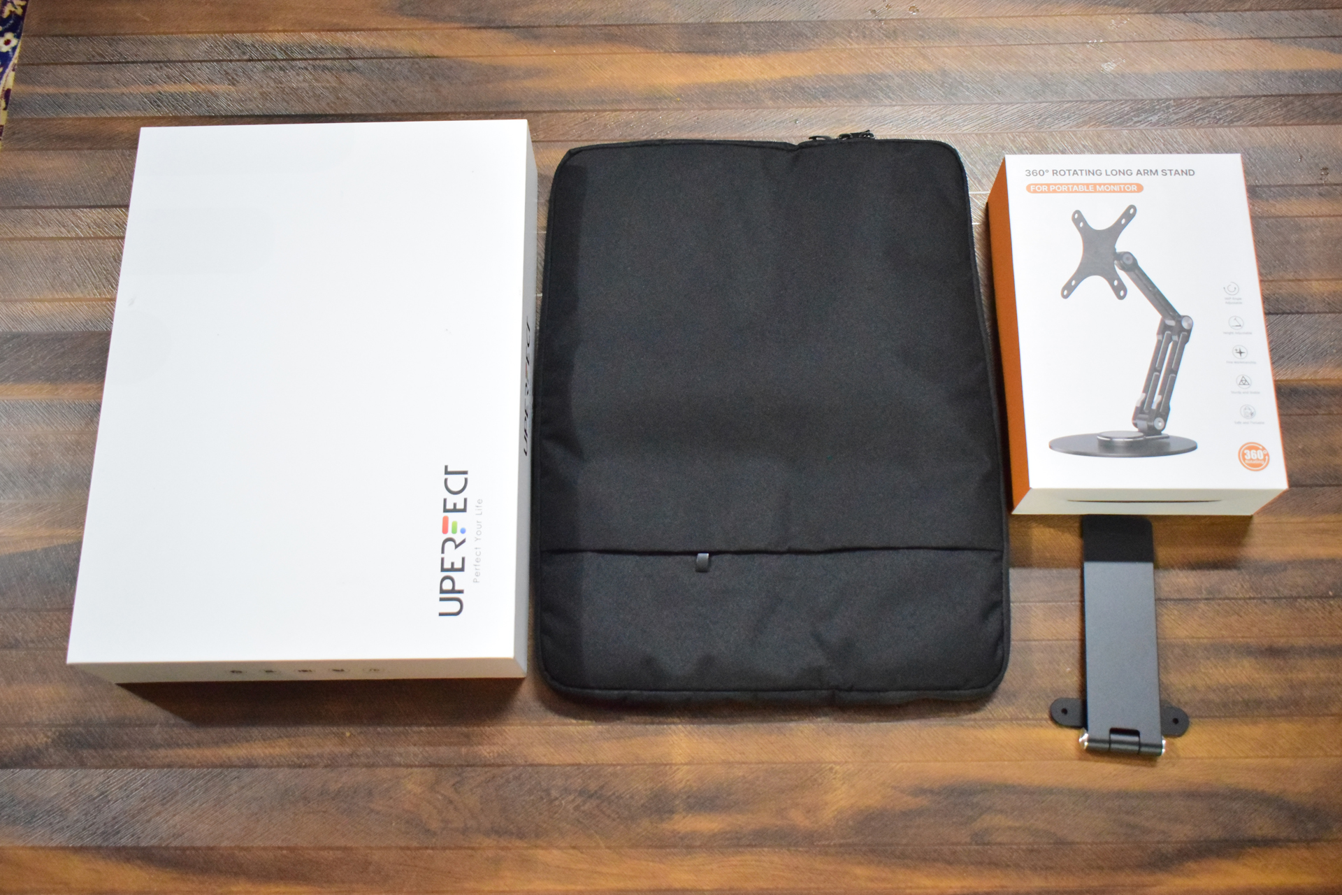 A white box labeled UPERFECT, a black protective case, and a box labeled 360° Rotating Long Arm Stand for Portable Monitor with the stand visible next to it are placed on a wooden surface.