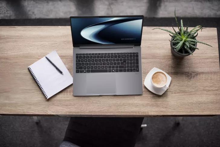 An ASUS ExpertBook laptop on a wooden desk alongside a notepad, pen, coffee cup, and potted plant.
